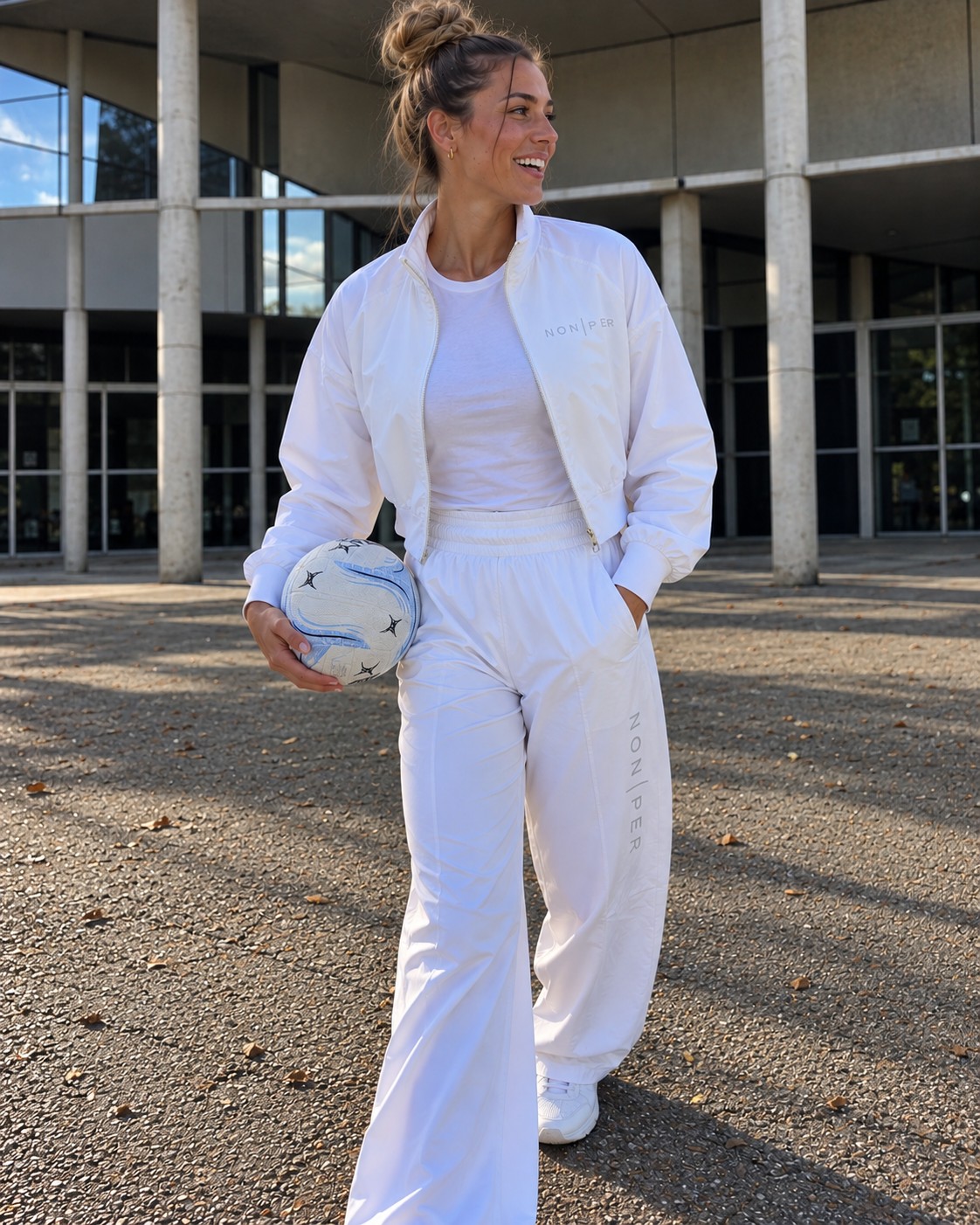 Smiling model walking outside a sports centre in the Line Jacket and Wide Leg, holding a netball